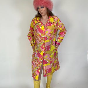 May include: A vintage-style, long-sleeved dress with a vibrant geometric pattern in yellow, pink, and white. The dress is buttoned down the front and paired with yellow boots and a pink fluffy hat. A pink bracelet is visible on the wrist.