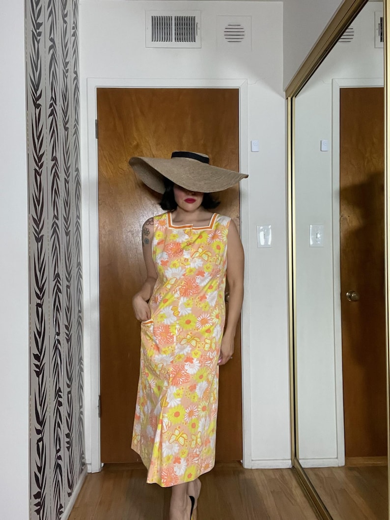 May include: A yellow, orange and white floral print midi dress with a square neckline and sleeveless design. The dress is paired with a wide, brown, floppy hat with a black band.