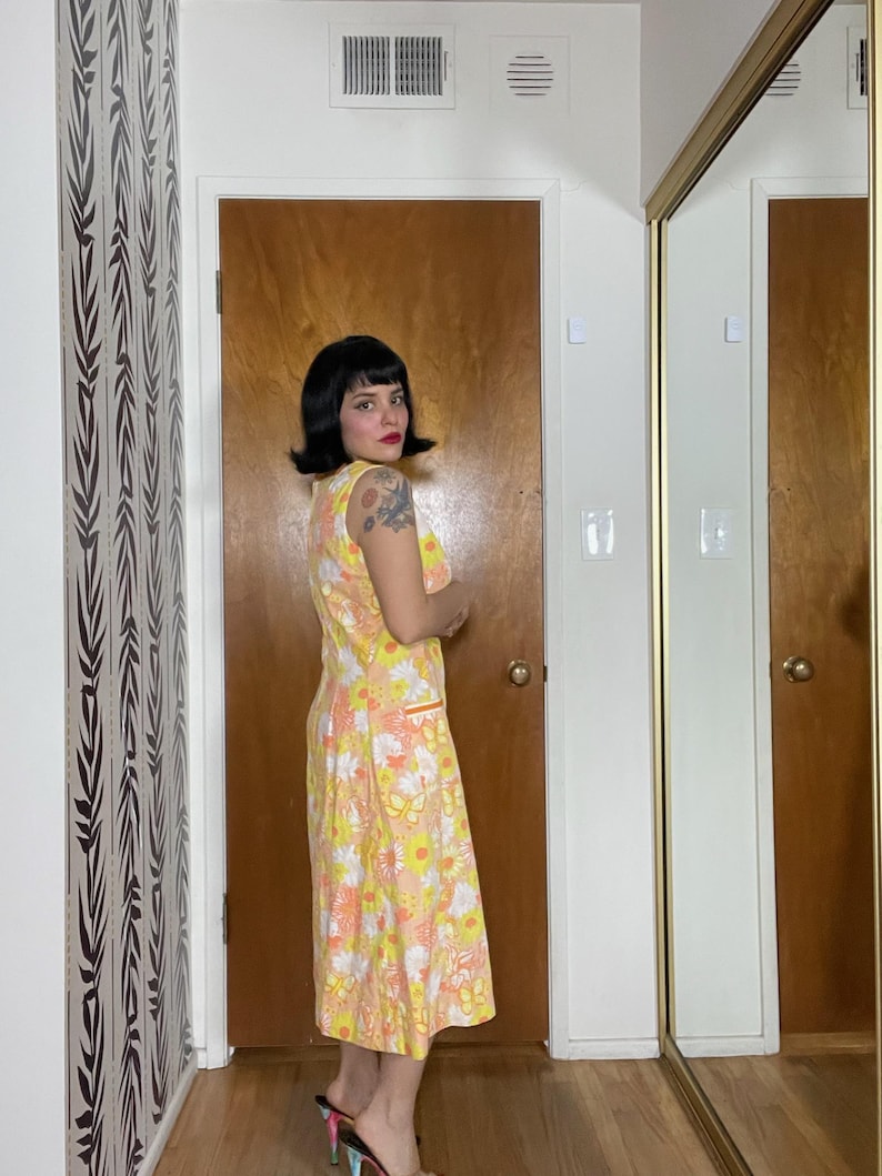 May include: A woman wearing a yellow, orange and white floral print dress with a sleeveless design. The dress has a fitted bodice and a flowing skirt. The woman is standing in a room with a wooden door and a mirror.
