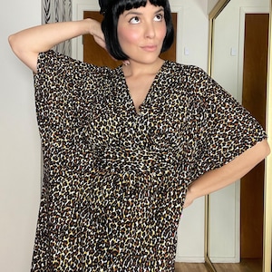 Puede incluir: Un vestido con estampado de leopardo con cuello en V. El vestido presenta un patrón de manchas marrones y negras sobre un fondo beige. La modelo lleva un sombrero negro con un adorno decorativo.