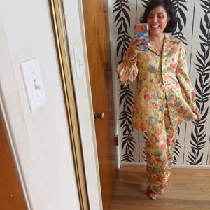 Puede incluir: Un traje de pantalón vintage con estampado floral en tonos amarillos, rosas y azules. El traje presenta una chaqueta con cuello y botones con mangas acampanadas y pantalones de pernera ancha a juego. El conjunto está hecho de una tela brillante.