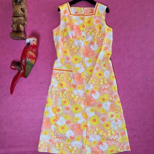 May include: A sleeveless dress with a floral print featuring butterflies, daisies, and other flowers in shades of yellow, orange, and pink. The dress has a square neckline and a button closure down the front.