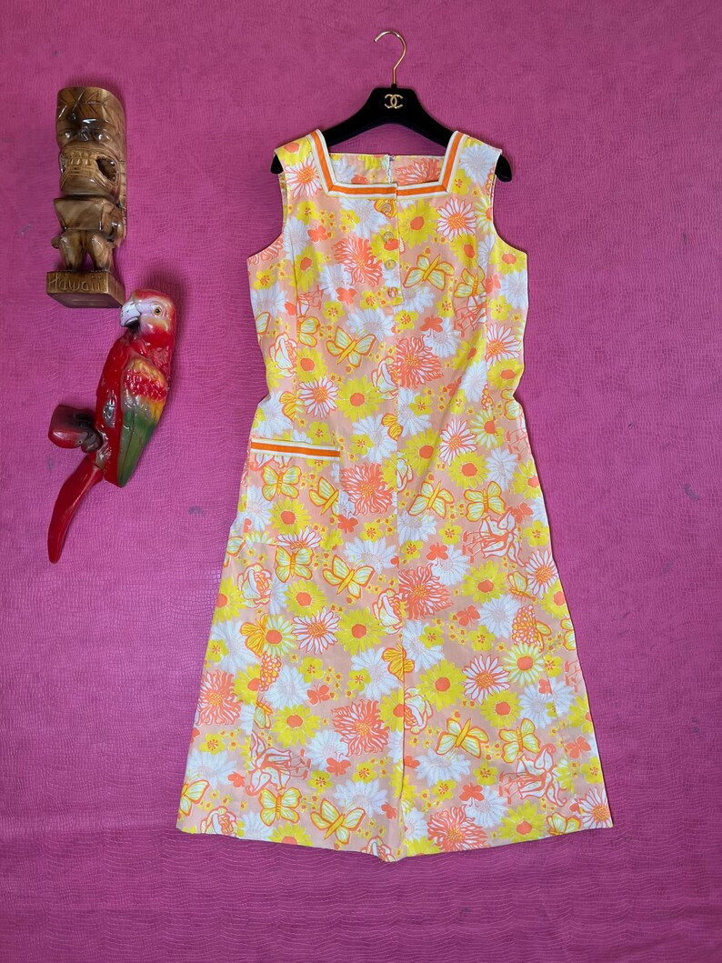 May include: A sleeveless dress with a floral print featuring butterflies, daisies, and other flowers in shades of yellow, orange, and pink. The dress has a square neckline and a button closure down the front.
