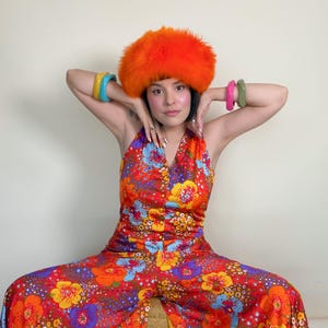May include: A vibrant, halter-neck jumpsuit with a bold floral pattern in red, orange, blue, and yellow. The outfit is paired with an orange faux fur hat and colorful bangles. The wide-leg design and bright colors evoke a retro, 70s-inspired aesthetic.