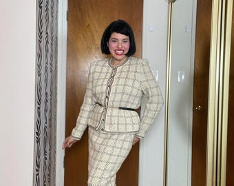 May include: A woman wearing a white and black checkered skirt suit with a black belt. She is standing in a room with a wooden door and a mirror.