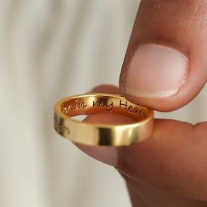 May include: A polished gold-tone band ring featuring a smooth, flat exterior surface. The inner circumference of the ring is engraved with the cursive text "ever in my Heart," visible along the interior edge of the metallic band.