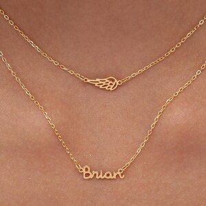 May include: A layered gold necklace with two chains. The top chain features a small wing pendant, while the bottom chain displays the name "Brian" in cursive script. The jewelry is displayed against a skin tone background.