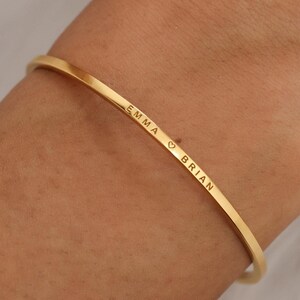 May include: A gold-colored bangle bracelet with the names "EMMA" and "BRIAN" engraved on it, separated by a heart symbol. The bracelet is thin and curved, designed to be worn on the wrist. The background is a soft, blurred white.