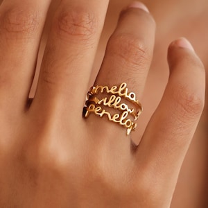 May include: A gold-colored ring with three bands, each bearing a name in cursive script: Amelia, Willow, and Penelope. The ring is worn on a finger, showcasing the personalized design. The ring is a piece of jewelry.
