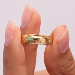 May include: A gold-colored ring with engraved script and a small, clear gemstone. The ring is held between fingers against a soft, neutral background. The ring's band is smooth and polished.