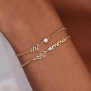 May include: Two gold bracelets are worn on a wrist. One bracelet features a small diamond and the name "emma" in cursive. The other bracelet has a wing charm. The bracelets are delicate and elegant.