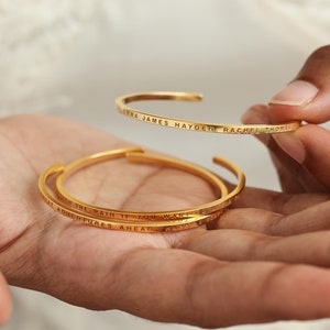 May include: Two gold bangle bracelets with engraved text. The top bracelet reads "EMMA JAMES HAYDEN RACHEL THOMAS". The bottom bracelet reads "BRUSH THE RAIN IF YOU WANT TO SEE GREAT ADVENTURES AHEAD HAPPY 5 YEARS".