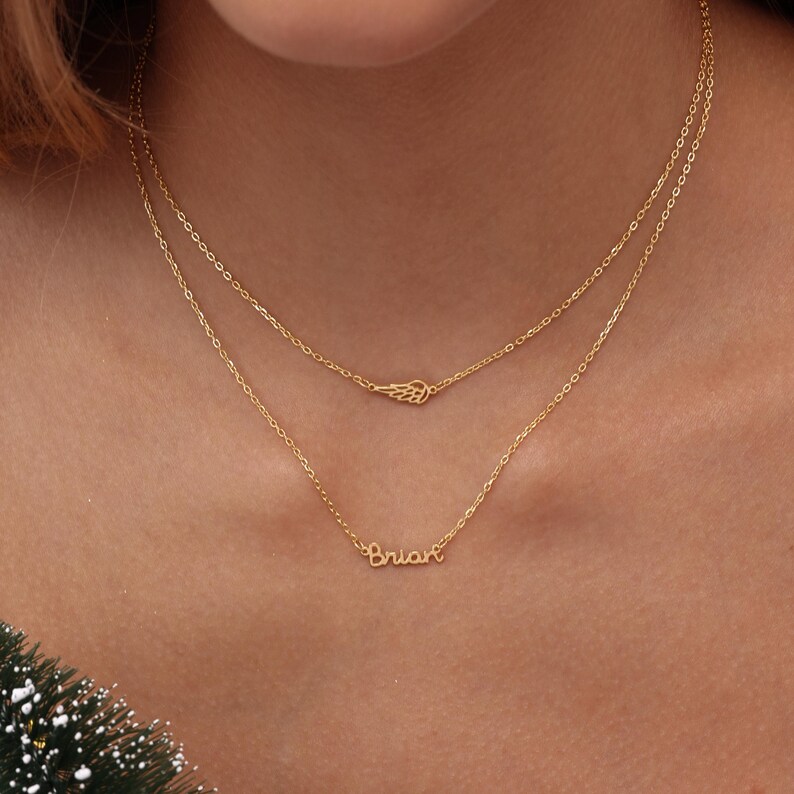 May include: Two delicate gold necklaces. The top necklace features a small wing pendant. The bottom necklace has the name "Brian" in cursive script. Both necklaces are layered and worn around the neck.