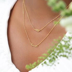 May include: A layered gold necklace featuring a delicate wing charm and a pendant spelling "Brian". The necklace is displayed against a soft, blurred background, highlighting the jewelry's intricate design and warm gold color.