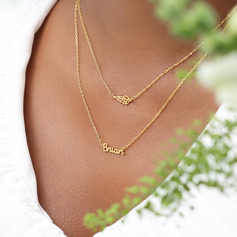 May include: A layered gold necklace featuring a delicate wing charm and a pendant spelling "Brian". The necklace is displayed against a soft, blurred background, highlighting the jewelry's intricate design and warm gold color.