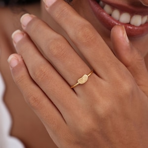 May include: A gold-colored ring featuring a fingerprint design. The ring has a thin band and a small, textured fingerprint shape at the center. The ring is worn on a finger.