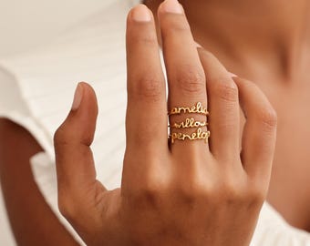 Puede incluir: Un conjunto de tres anillos dorados apilados en un dedo, cada uno con un nombre diferente escrito en una delicada letra cursiva. Los nombres que aparecen en los anillos de arriba a abajo son Amelia, Willow y Penelope en alambre fino.