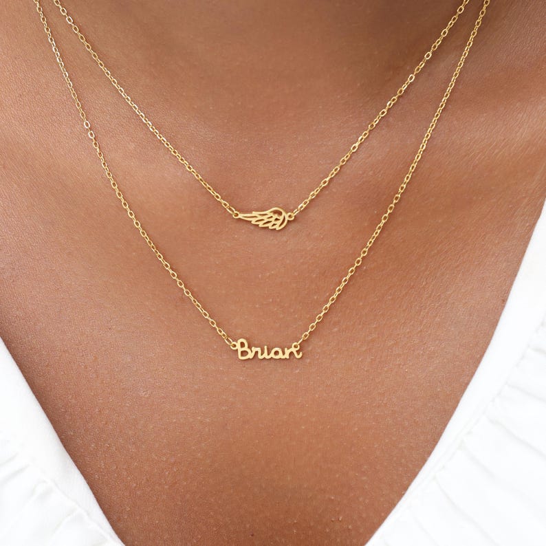 May include: A layered gold necklace featuring a delicate wing charm and a cursive name pendant that reads "Brian". The necklace is displayed against a white background.