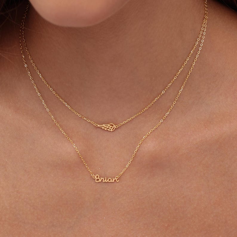 May include: A layered gold necklace with two chains. The top chain features a small wing pendant, while the bottom chain displays the name "Brian" in cursive script. The jewelry is displayed against a skin tone background.