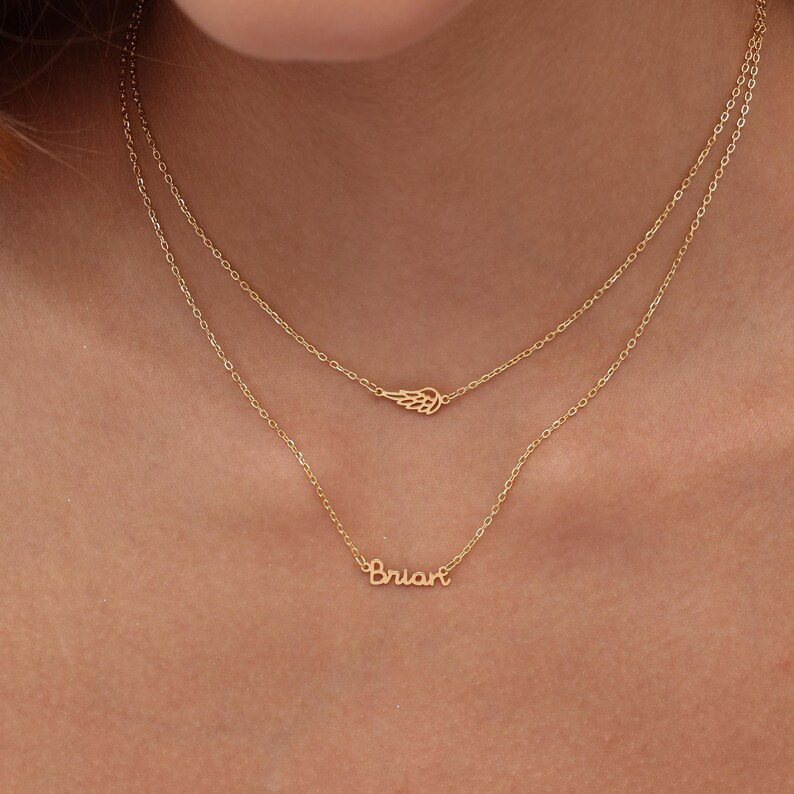 May include: A layered gold necklace set. The top chain features a small wing pendant, while the lower chain displays the name "Brian" in cursive script. The necklaces are delicate and appear to be made of gold.