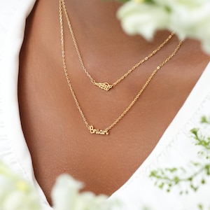 May include: A layered gold necklace with two chains. The top chain features a small wing charm, while the lower chain spells out the name "Brian" in cursive script. The necklace is worn against a white shirt.