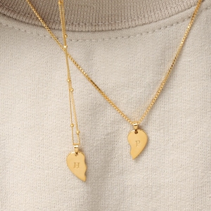 May include: Two gold-toned necklaces featuring heart-shaped pendants split into halves. One pendant is engraved with the letter H, and the other is engraved with the letter P. The necklaces have delicate chains with small bead accents.