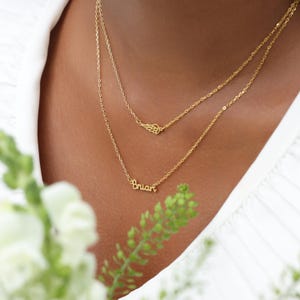May include: Two gold necklaces layered on a person. The top necklace features a small wing charm, while the bottom necklace spells out the name "Brian" in cursive. The necklaces are delicate and elegant, perfect for everyday wear.
