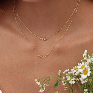 May include: A layered gold necklace featuring a delicate wing pendant and a script nameplate that reads "Brian". The necklace is shown against a skin tone background, with white flowers in the lower right corner.