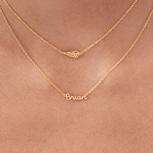 May include: Two gold necklaces. The top necklace features a small wing charm. The bottom necklace spells out the name "Brian" in cursive script. Both necklaces are delicate and appear to be made of gold.