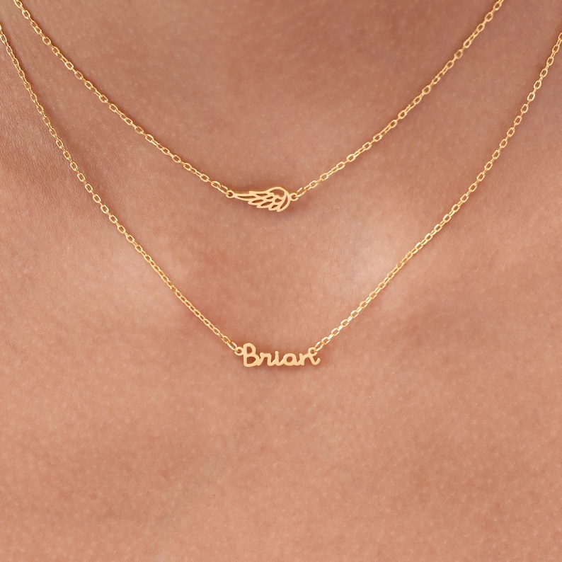 May include: Two gold necklaces. The top necklace features a small wing charm. The bottom necklace spells out the name "Brian" in cursive script. Both necklaces are delicate and appear to be made of gold.