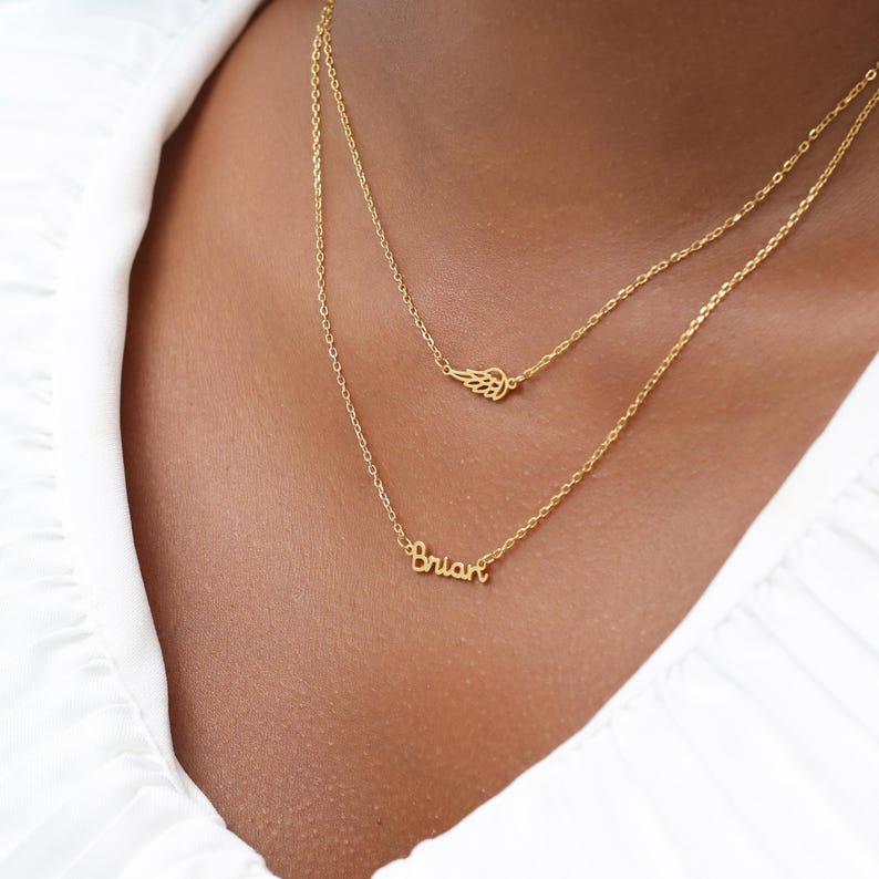 May include: A layered gold necklace with two chains. The top chain features a small wing charm, while the bottom chain displays the name "Brian" in a delicate script. The necklace is worn against a white shirt.