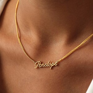 May include: A gold-colored necklace with the name "Penelope" in cursive script. The pendant is attached to a delicate chain. The necklace is worn around the neck, against a skin tone background.