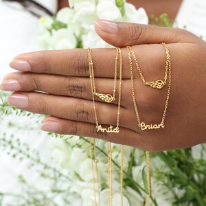 May include: Gold-colored necklaces with delicate chains and personalized name pendants. Each necklace features a small wing charm. One necklace reads "Anita", and another reads "Brian". The necklaces are displayed against a backdrop of white flowers.