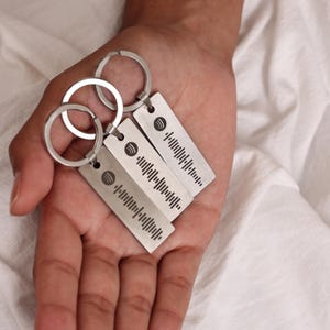 May include: Three silver keychains with a Spotify logo and a barcode-like design. The keychains are held in a hand.