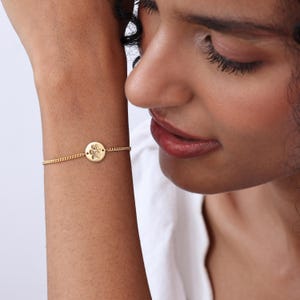 May include: A gold-toned chain bracelet featuring a small, circular charm engraved with a detailed paw print design. The delicate metal chain connects to the center disc, which displays a textured paw impression on its polished surface.