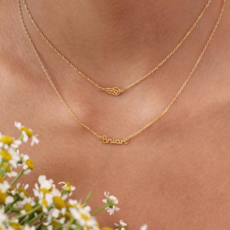 May include: Two delicate gold necklaces. The top necklace features a small wing charm. The bottom necklace has the name "Brian" in cursive script. The necklaces are layered and worn close to the neck.