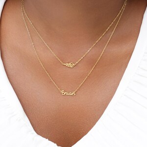 May include: A layered gold necklace set. The top chain features a small wing pendant, while the lower chain spells out the name "Brian" in cursive script. The necklaces are displayed against a white background.