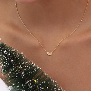 May include: A delicate gold necklace with a small, curved pendant. The necklace features a fine chain with tiny beaded accents. The pendant has engraved text. The necklace is worn on a person.