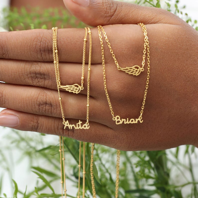May include: Gold-colored necklaces with delicate chains and personalized name pendants. Each necklace features a small wing charm. One necklace says "Anita" and another says "Brian". The necklaces are displayed on a hand.
