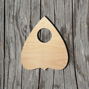 May include: A wooden planchette, a tool used in Ouija board games, shaped like a heart with a circular hole in the center. The planchette is made of light wood and is sitting on a weathered wooden surface.