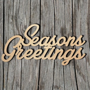 May include: Wooden sign with the text "Seasons Greetings" in a cursive font. The sign is light brown and is placed on a weathered wood background.
