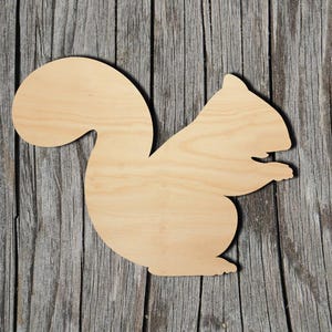 May include: A flat, light-colored wooden cutout in the shape of a squirrel. The silhouette features a large, curved tail, a rounded body, and a head with a small pointed ear, showing the natural grain of the wood across the entire surface.