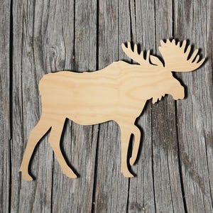 May include: A flat, light-colored wooden cutout in the shape of a moose. The silhouette features the distinct profile of the animal, including its large antlers, a prominent hump on the back, and four legs positioned as if the moose is walking.