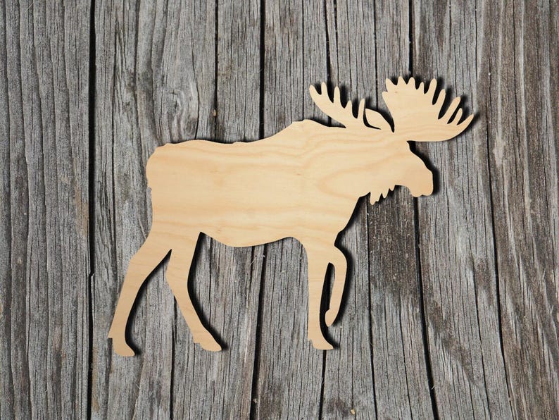 May include: A flat, light-colored wooden cutout in the shape of a moose. The silhouette features the distinct profile of the animal, including its large antlers, a prominent hump on the back, and four legs positioned as if the moose is walking.