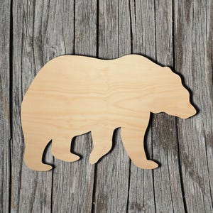 Pode incluir: Um recorte de madeira de cor clara em forma de urso. O objeto apresenta uma superf&iacute;cie lisa com veios de madeira natural e uma silhueta distinta que mostra a cabe&ccedil;a, as costas e as quatro patas do animal em posi&ccedil;&atilde;o de perfil.
