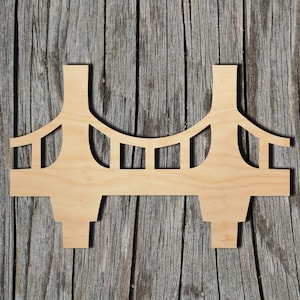 May include: A wooden cutout of a bridge, featuring two towers connected by a curved top and a series of vertical supports. The bridge is made of light-colored wood and is set against a weathered, gray wooden background. The design is simple and clean.