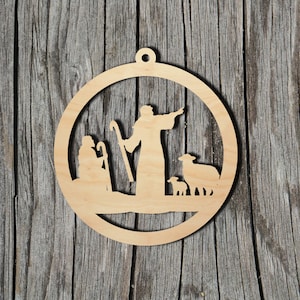 Könnte beinhalten: Ein kreisförmiges Ornament aus hellem Holz mit einer lasergeschnittenen Silhouette einer Krippenszene. Das Design zeigt zwei Figuren mit Stäben, von denen eine steht und in eine Richtung zeigt, neben einem kleinen Lamm und einem Schaf.