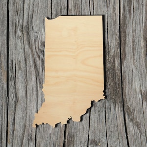 May include: A wooden cutout of the state of Indiana. The cutout is light brown and has a smooth surface. It is placed on a weathered wooden background.