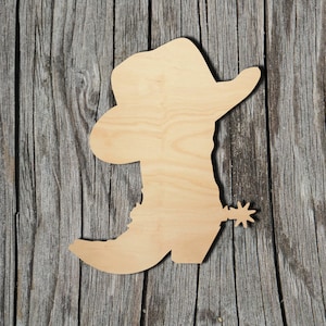May include: Wooden cutout of a cowboy boot with a hat on top. The boot has a spur on the heel. The cutout is made of light brown wood and is on a gray wooden background.