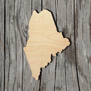 May include: A wooden cutout of the state of Maine. The cutout is light brown and has a smooth, unfinished surface. It is placed on a weathered wooden background.